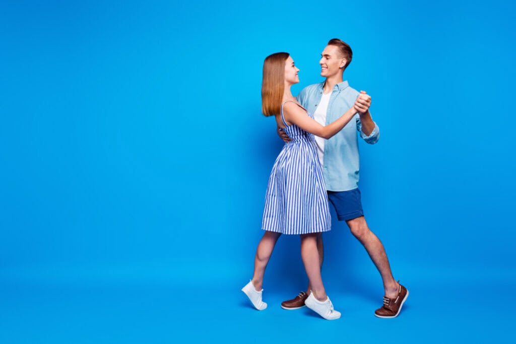 A couple dances together against a solid blue background. The woman wears a striped dress and sneakers. The man wears a light shirt, blue shorts, and sneakers.