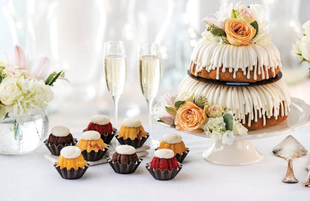 A decorated layer cake with white icing and flowers, mini bundt cakes with icing, two champagne glasses, and white floral arrangements on a white tablecloth.