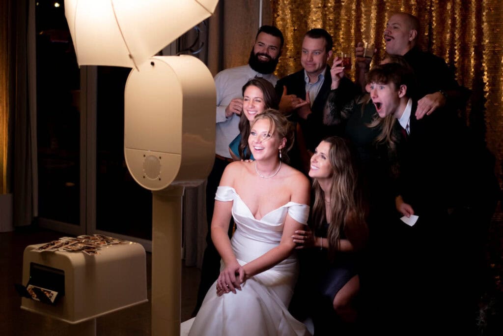 A bride sits in front of a photo booth, surrounded by friends posing and smiling against a gold sequin backdrop at a celebration.