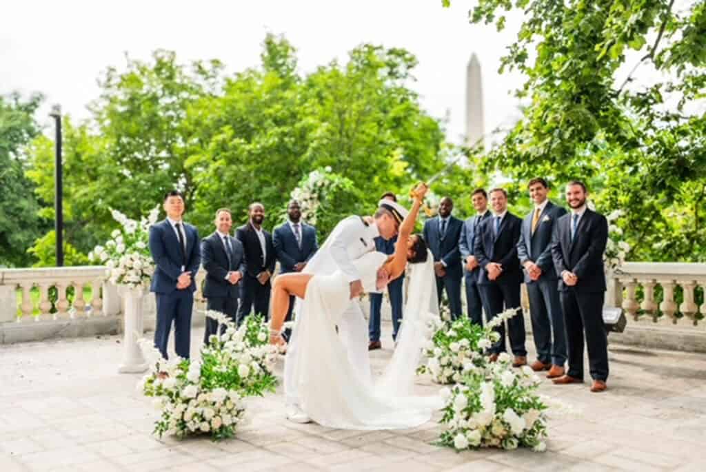 A bride and groom kiss while dancing outdoors, surrounded by floral arrangements and groomsmen in suits, with trees and a monument in the background.