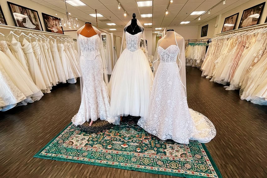 Three wedding dresses are displayed on mannequins in the center of a bridal shop, with more dresses hanging along the walls on racks.