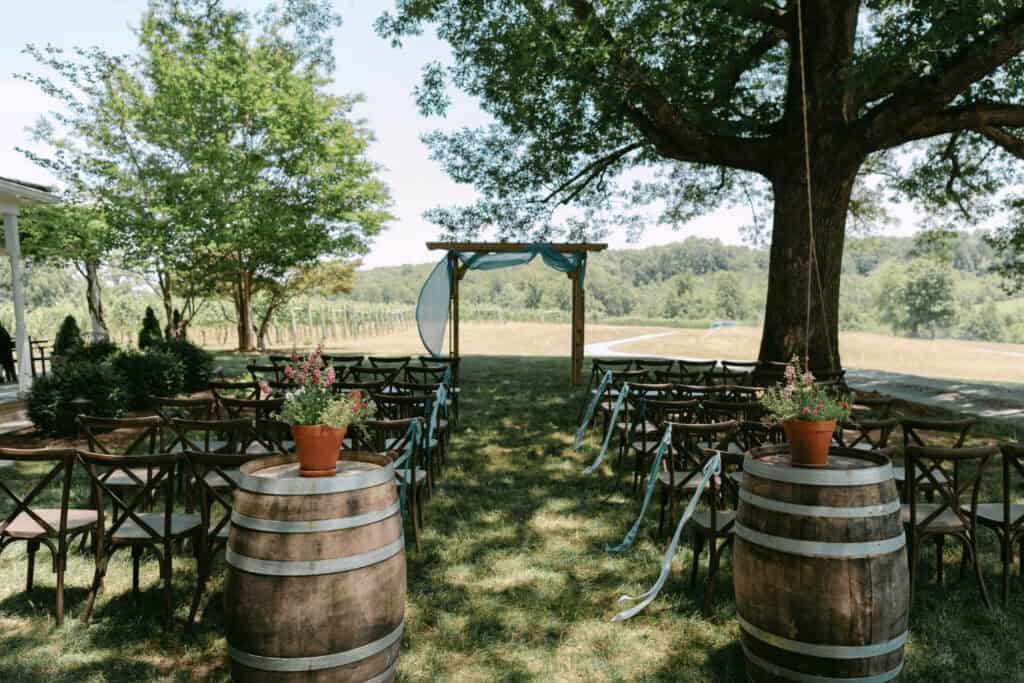 Outdoor wedding ceremony setup with wooden chairs facing an arbor, two decorated barrels in the foreground, and trees and fields in the background.