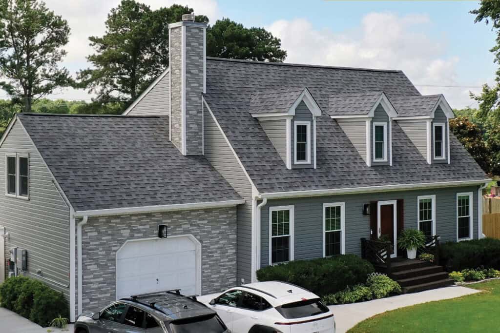 A two-story gray house with a gable roof, dormer windows, a brick chimney, attached garage, and two cars parked in the driveway.
