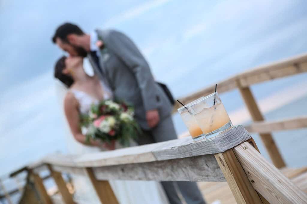 A close-up of a cocktail glass on a wooden railing with a bride and groom kissing in the blurred background outdoors.