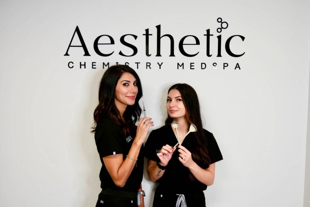 Two women in black uniforms stand in front of an Aesthetic Chemistry Med Spa sign, holding cosmetic tools and smiling at the camera.