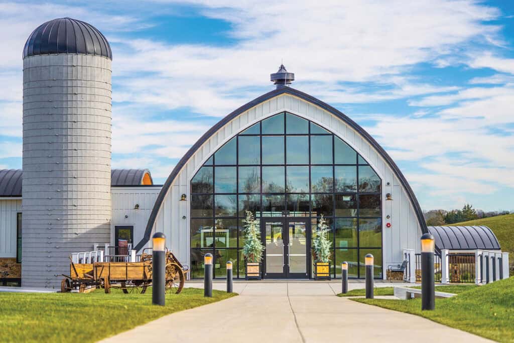 A modern barn-style building with a large arched glass entrance, adjacent to a tall silo, set on a grassy landscape under a blue sky.
