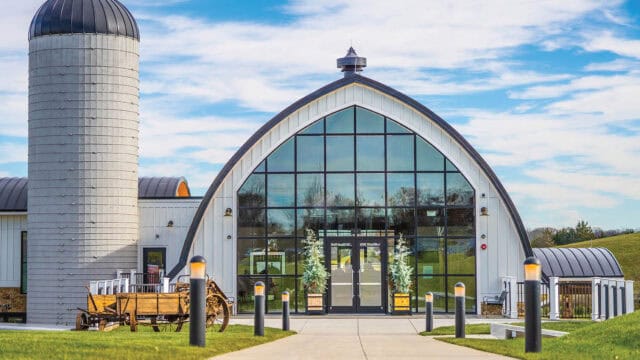 A modern barn-style building with a large arched glass entrance, adjacent to a tall silo, set on a grassy landscape under a blue sky.