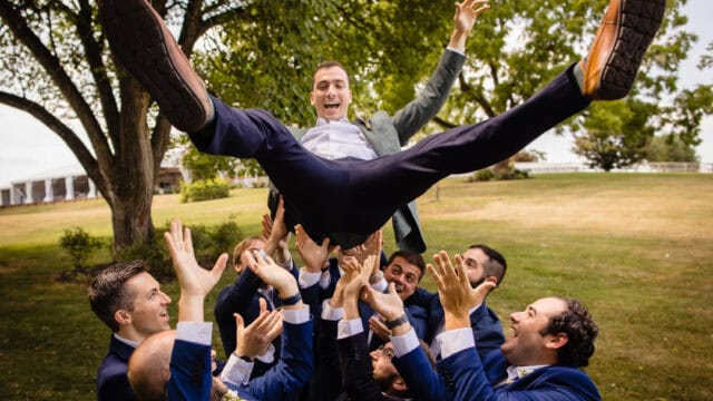 A group of men in suits lift and toss another man into the air outdoors, with smiles and trees in the background.
