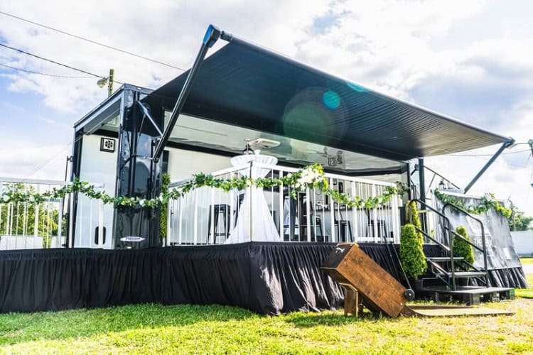 A portable stage setup with black awning, white table and chairs, and green garland decorations, placed outdoors on grass under a partly cloudy sky.