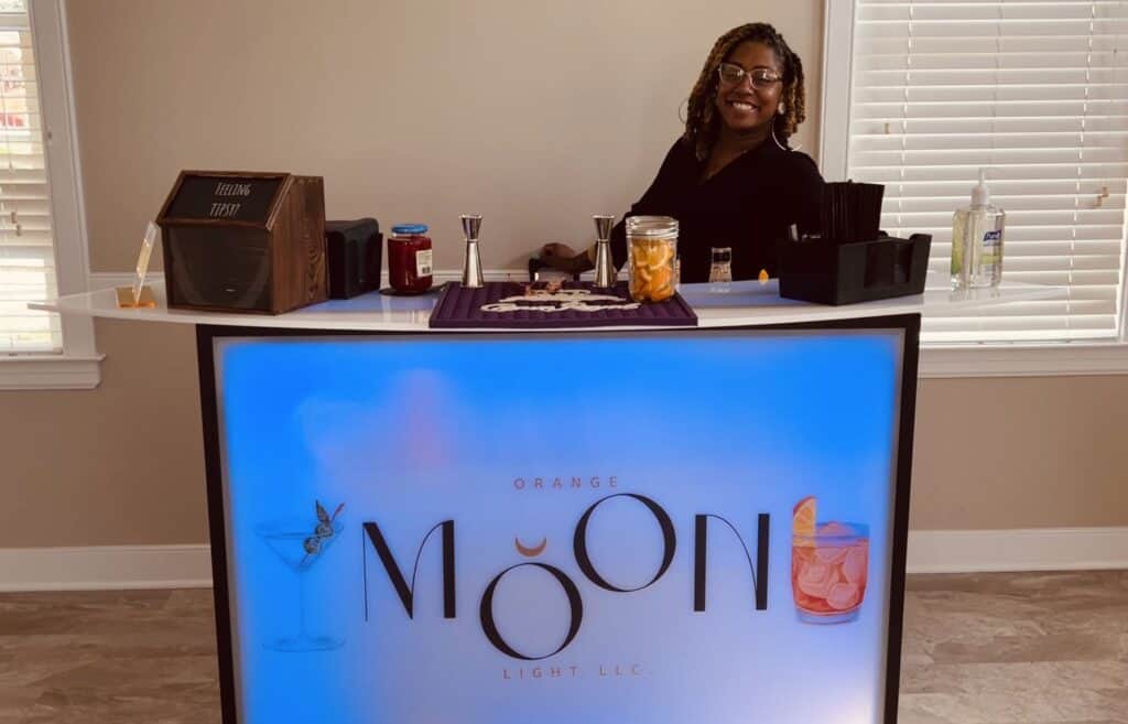 A woman stands behind a lit bar counter labeled Orange Moon Light LLC, with cocktail ingredients and tools arranged on the counter.