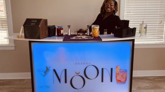 A woman stands behind a lit bar counter labeled Orange Moon Light LLC, with cocktail ingredients and tools arranged on the counter.
