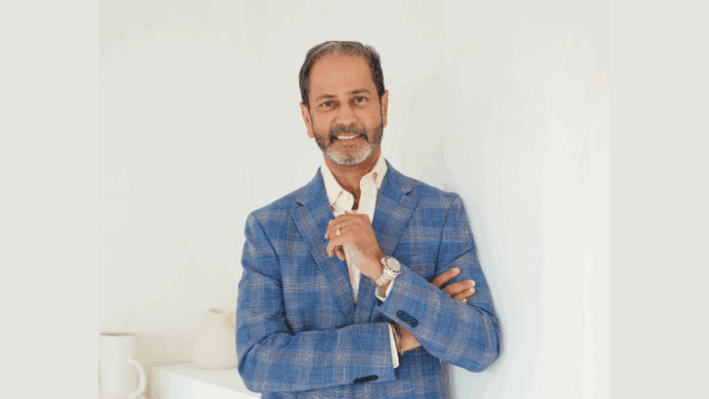 A man with short hair and a beard in a blue checked blazer stands with crossed arms in a bright, minimalist room with white decor.