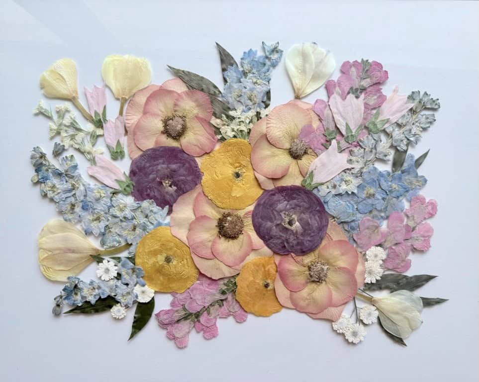 Pressed flower arrangement featuring yellow, purple, pink, and white blooms with green leaves displayed on a white background.