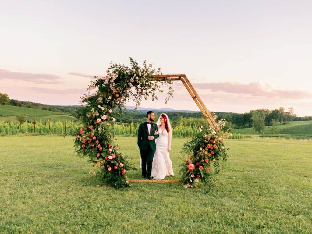 A bride and groom stand together inside a large hexagonal floral arch on a grassy field, with rolling hills and a sunset sky in the background.