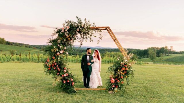 A bride and groom stand together inside a large hexagonal floral arch on a grassy field, with rolling hills and a sunset sky in the background.